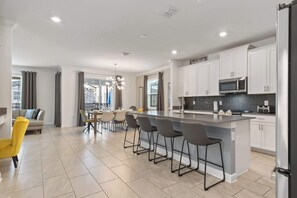 A modern kitchen with white cabinets, a central island with bar stools, a microwave, and a refrigerator. - Storey Lake 6BR Villa | Water Park + Pool Spa (Kissimmee)