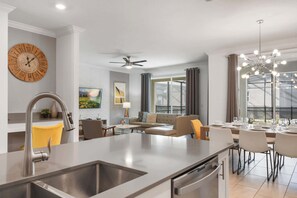 A modern kitchen with a central island, a dining area with a chandelier, and a living space with a sofa and a painting. - Storey Lake 6BR Villa | Water Park + Pool Spa (Kissimmee)