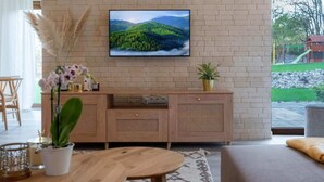 A living room with a TV mounted on a brick wall, a wooden cabinet with a plant, and a sofa. - Comfortable holiday villa, sauna, jacuzzi (Marcyporeba)
