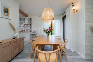 Uma área de refeições com mesa e cadeiras de madeira, um lustre e vista para o exterior pela janela.