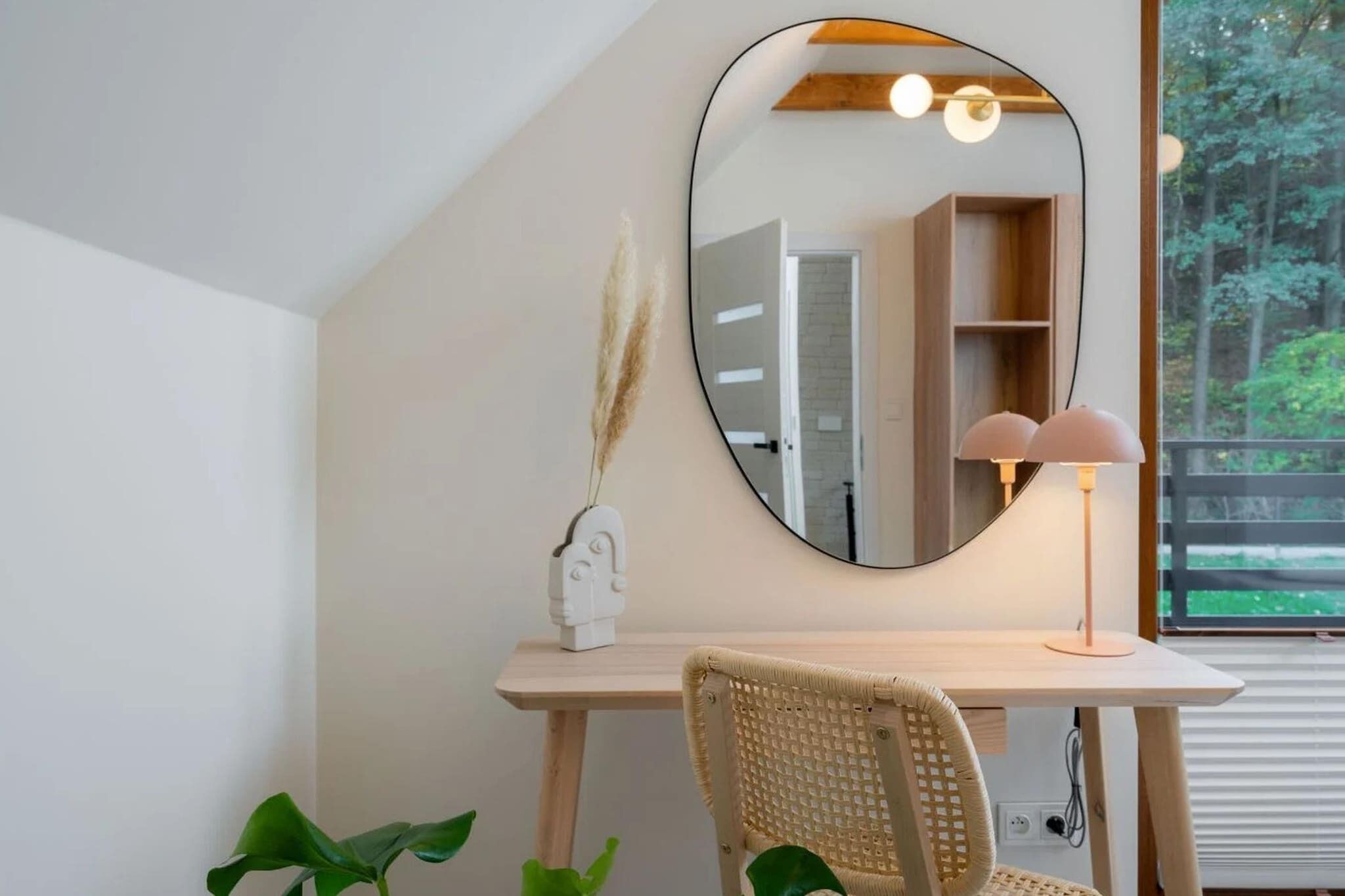 Een kamer met een bureau, een stoel, een spiegel, een lamp en een raam met uitzicht op bomen.