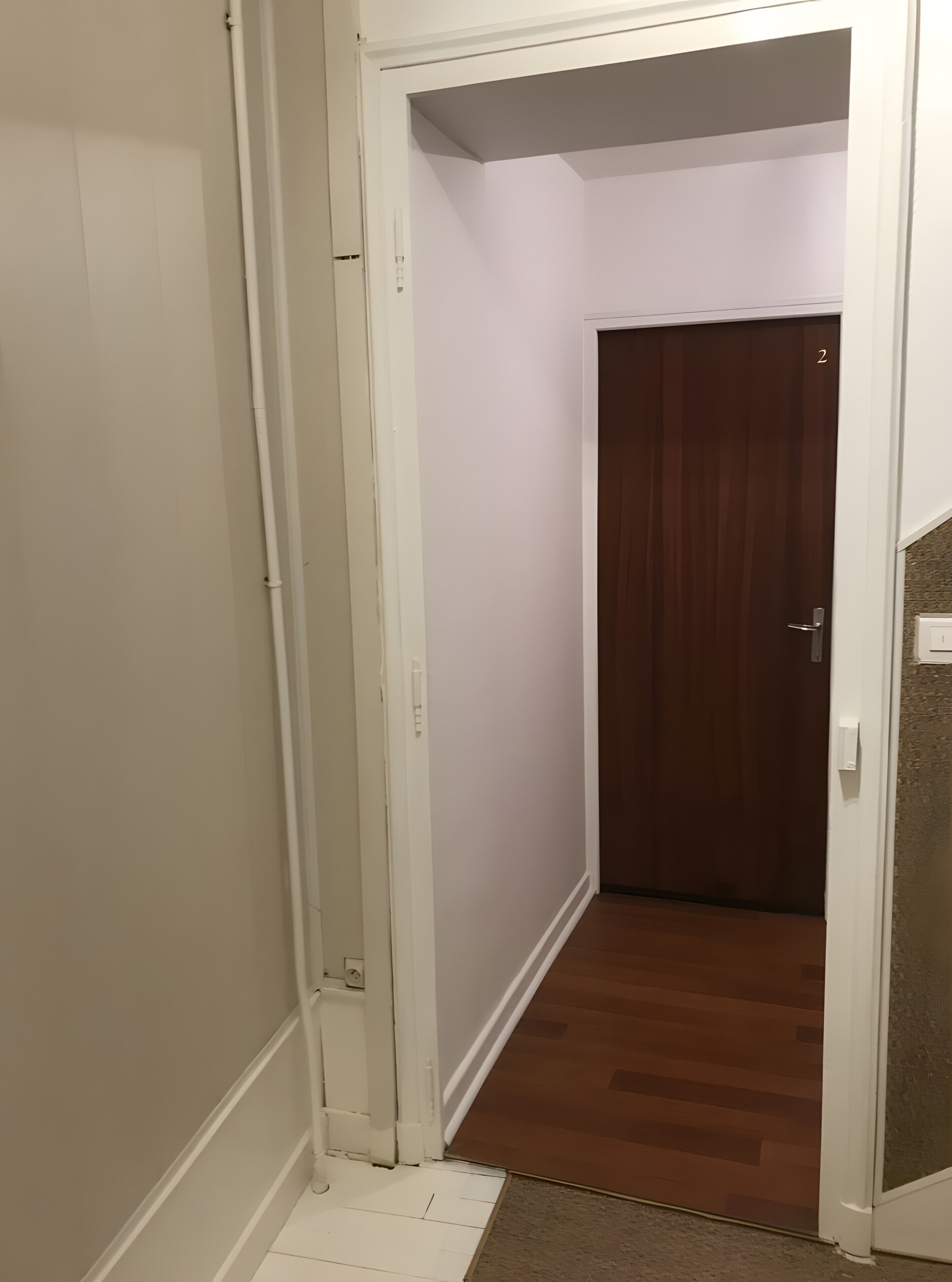 A narrow hallway with a wooden door at the end, white walls, and a white baseboard.