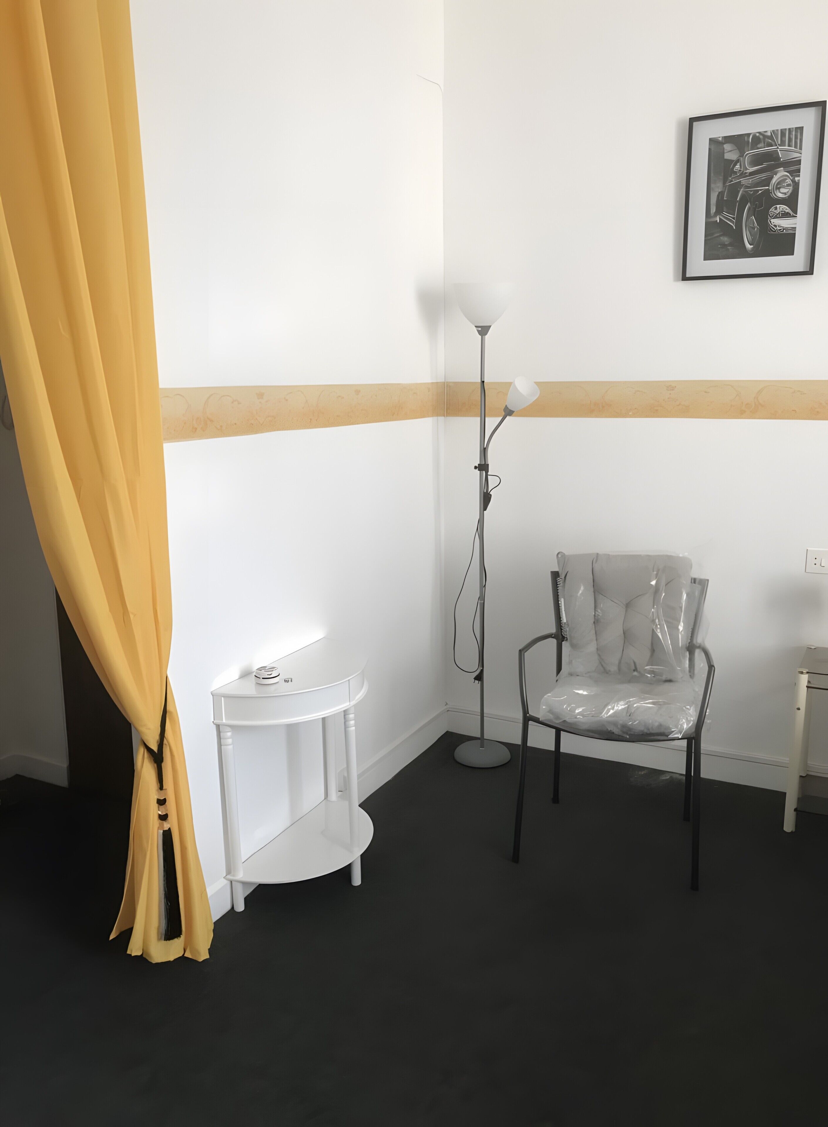 Quarto de canto com uma mesa branca, uma cadeira, um abajur e um quadro na parede.