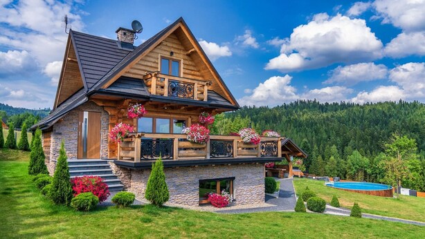 A two-story log cabin with a stone foundation, a balcony with flower boxes, and a swimming pool.