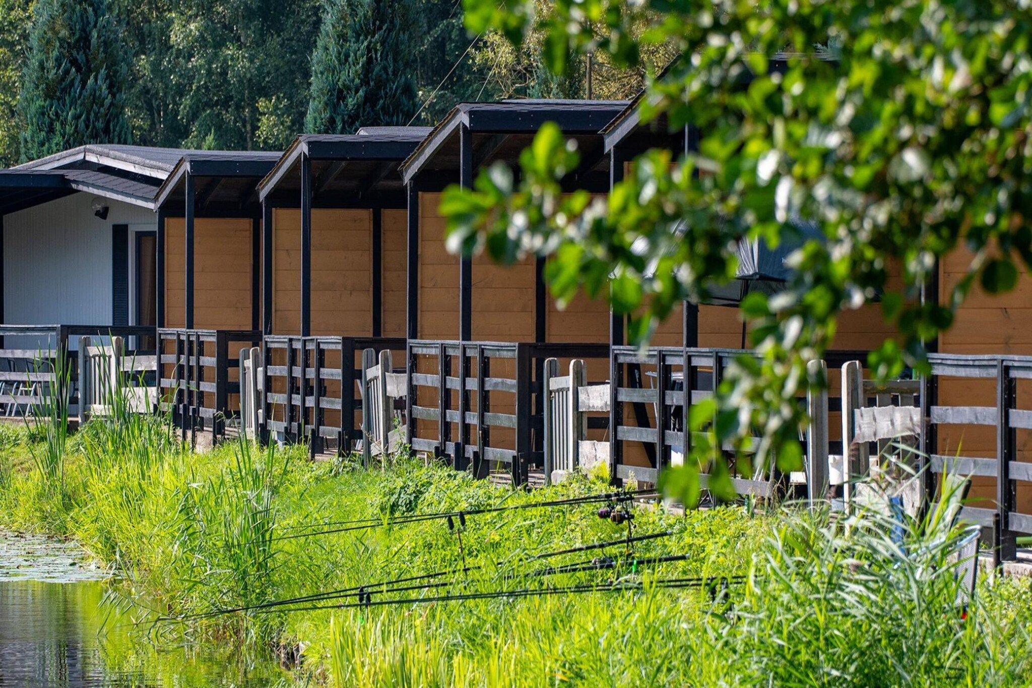 Une rangée de cabanes en bois, avec une rambarde en métal à l’avant, se dressant sur un fond de verdure et au bord d’une étendue d’eau.