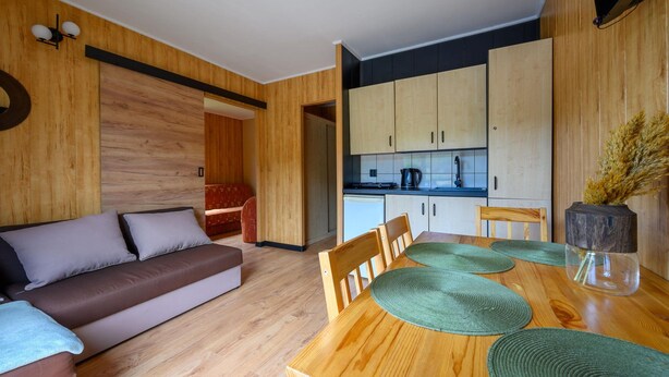 A modern living space with a wooden floor, a sofa, a dining table with chairs, and a kitchen area. - Waterfront Holiday Home in Darlowo (Darlowo)