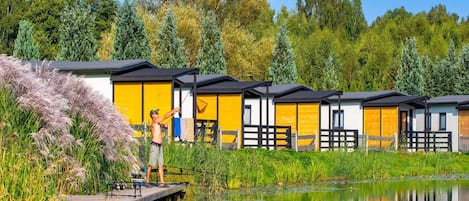 Een rij gele chalets met een steiger die uitsteekt in een kalm waterlichaam, omringd door weelderige begroeiing en bomen.