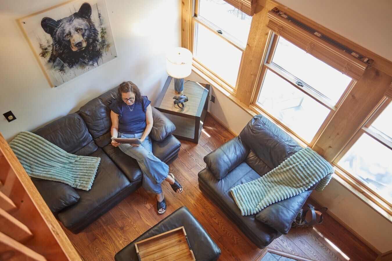 A cozy living room with a person sitting on a couch, a table with a lamp, and a large window with a view.