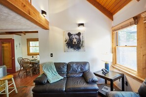 A cozy living room with a leather sofa, a dining area with wooden chairs, and a large window with blinds. - Black Bear Resort - Serene Cabin Retreat | Views, Deck & Resort Access (Davis)