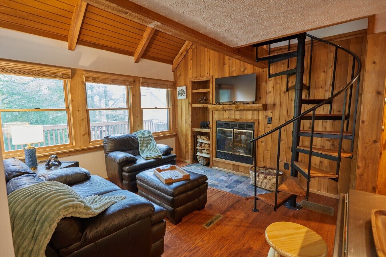 A cozy cabin interior with a fireplace, a sofa, a chair, and a wooden staircase.