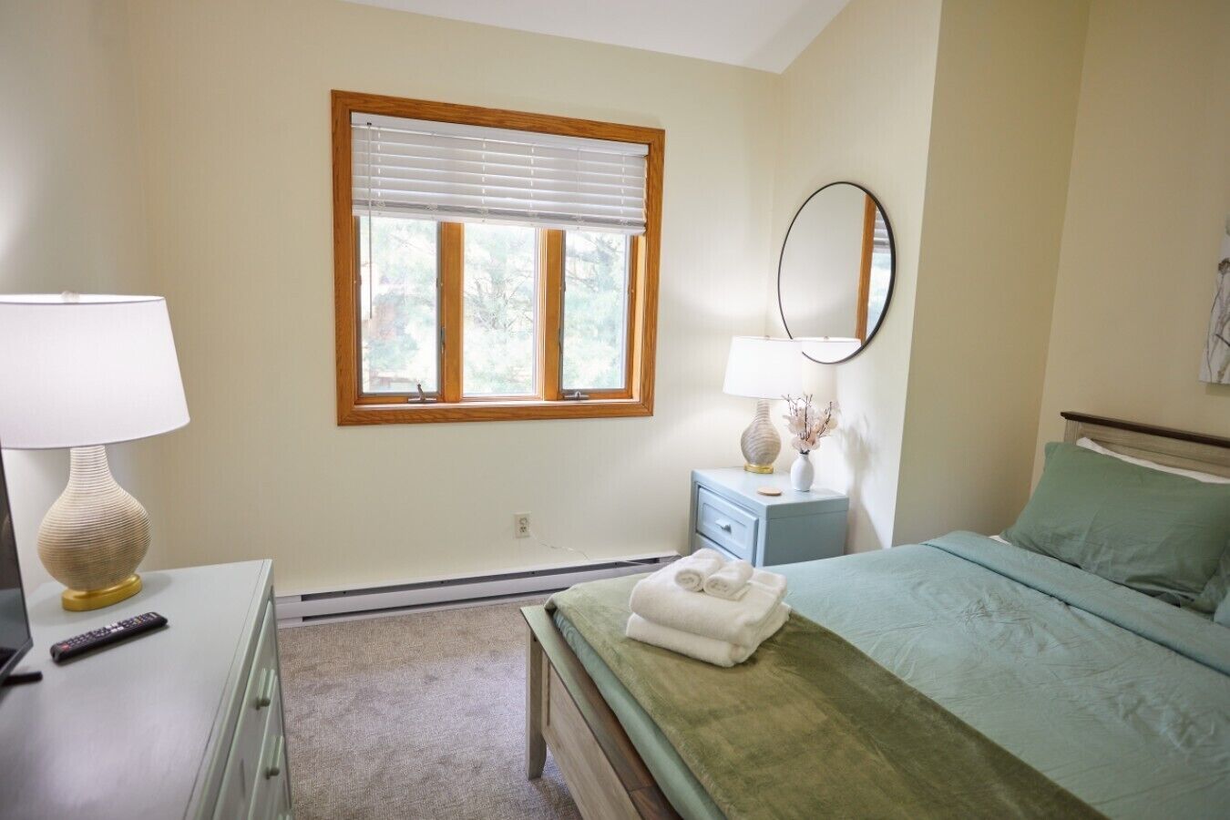 A bedroom with a bed, a bedside table with a lamp, a dresser, a window with blinds, and a mirror on the wall.