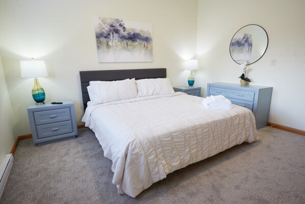 Une chambre à coucher comprenant un lit, deux tables de chevet, une commode, une lampe, un miroir et un tableau accroché au mur.