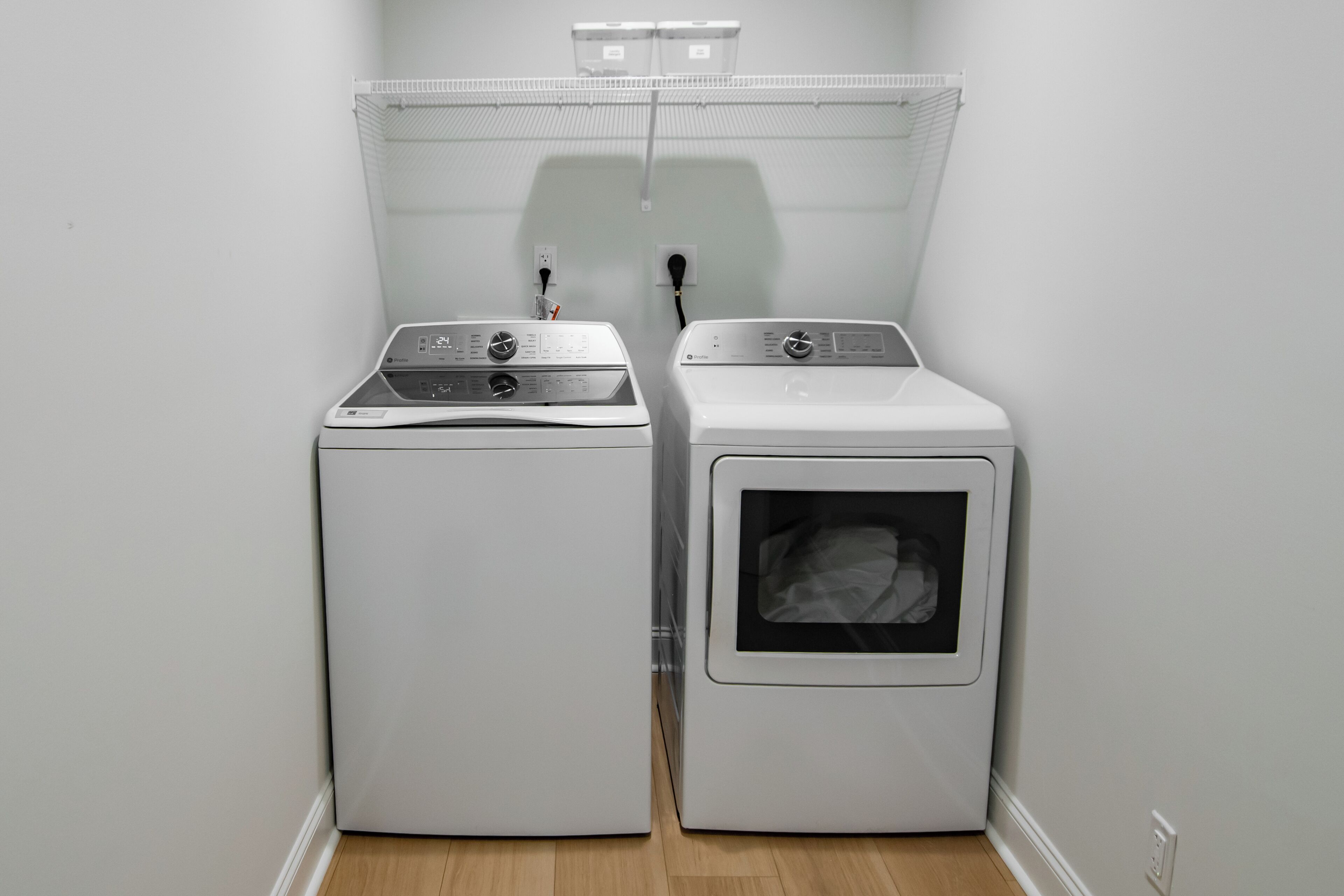 A laundry room with two white appliances, a wall-mounted shelf above, and a power outlet.