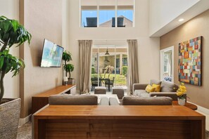Ein geräumiges Wohnzimmer mit Couchtisch aus Holz, grauen Sofas, einem an der Wand montierten Fernseher und einem großen Fenster mit Vorhängen.