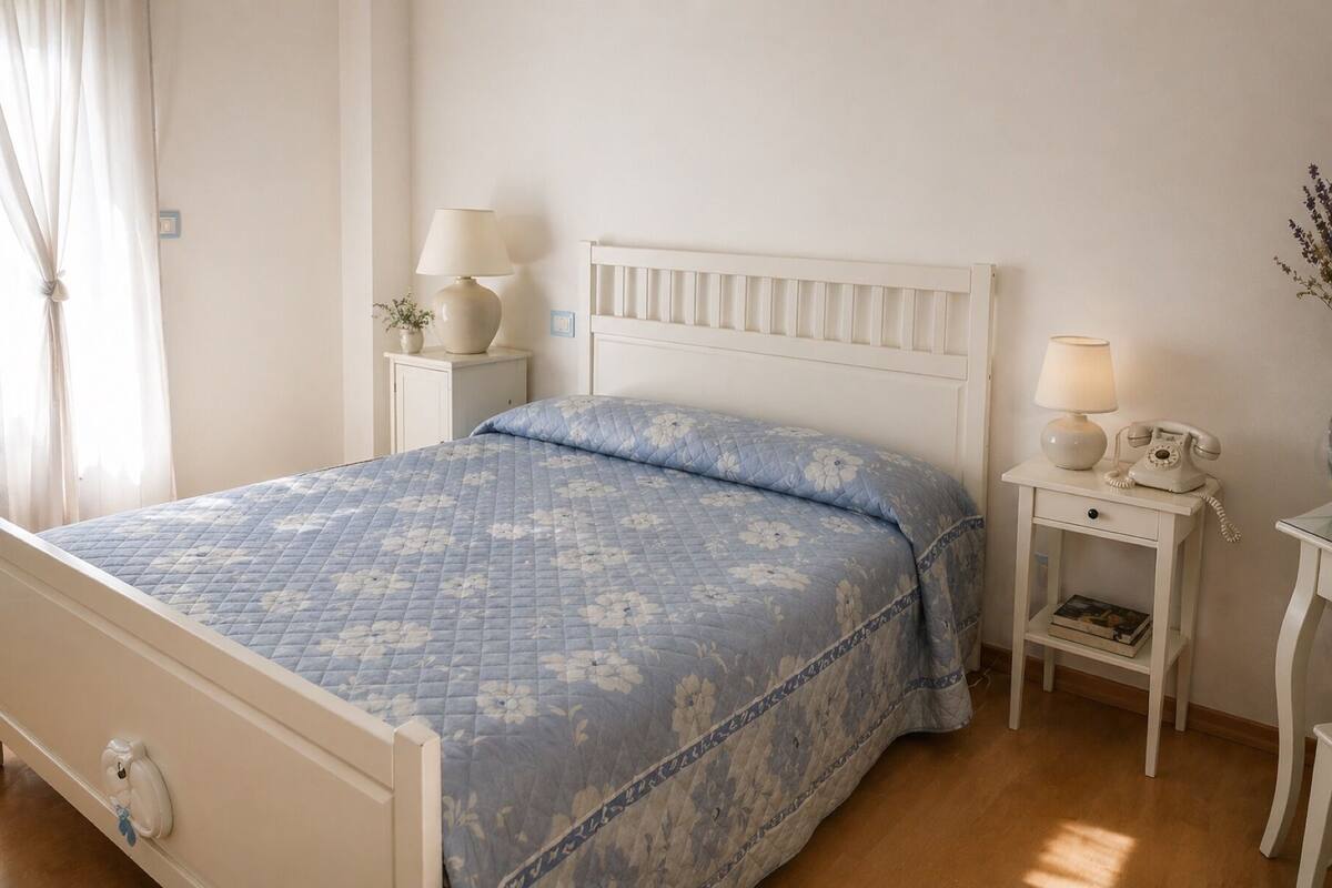 A bed with a floral quilt, bedside tables with lamps, a nightstand with a telephone, and a small shelf with a book.