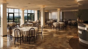 Un vaste espace dédié à la salle à manger, avec plusieurs tables et chaises rondes, de grandes fenêtres offrant une vue dégagée et un plancher ciré.