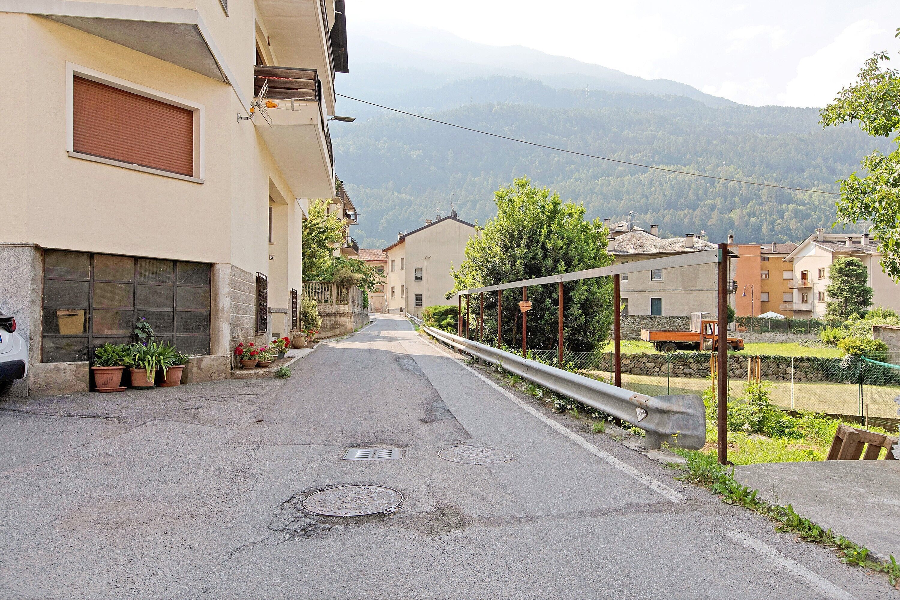Une rue étroite, bordée de bâtiments des deux côtés, avec une barrière métallique et une montagne en arrière-plan.