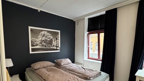 Ein Schlafzimmer mit einem Bett, einem Nachttisch, einem Wandbild, einem Fenster mit Vorhängen und einer Deckenleuchte.