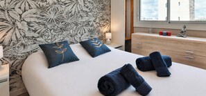 Ein ordentlich bezogenes Bett mit zwei Kissen, ein Nachttisch mit Lampe, eine hölzerne Kommode und ein Fenster mit Blick auf Gebäude.