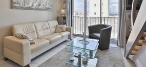 Ein modernes Wohnzimmer mit einer Couch, einem Glastisch und Blick auf die Stadt durch große Fenster.