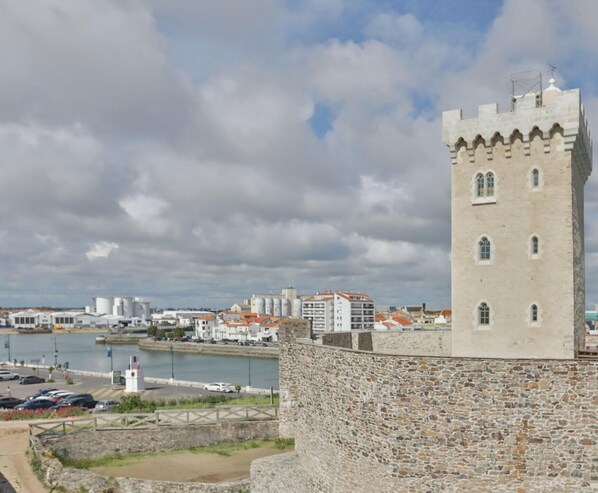 Una torre in pietra con merli, un corpo d'acqua e edifici sullo sfondo.