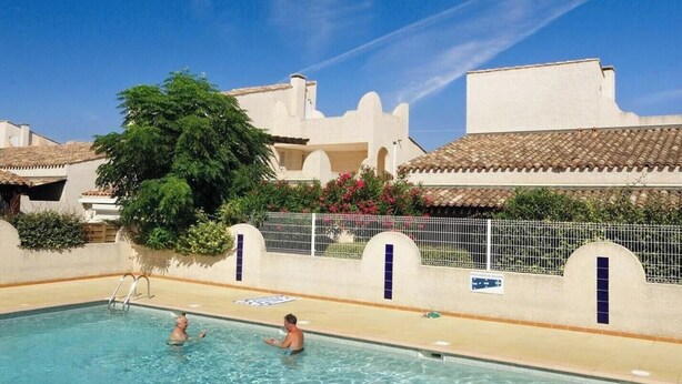 A clear swimming pool with two people in the water, surrounded by white buildings with terracotta roofs and a tree. - Beach Holiday Home in Cap D'agde (Agde)