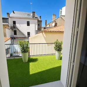Ein Balkon mit Kunstgrasanlage, zwei Blumentöpfen, einem Metallgeländer und Blick auf benachbarte Gebäude.