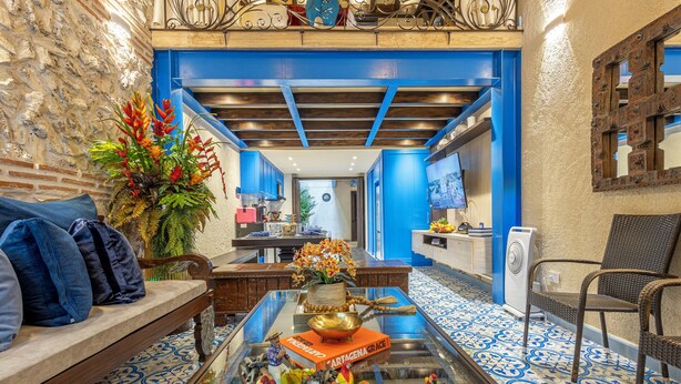Un hall d’hôtel avec un sol carrelé de bleu et de blanc, un banc en bois avec des coussins bleus, une table en verre surmontée d’un panier de fruits et d’un livre intitulé « Cartagena Grace », ainsi qu’une plante décorative.