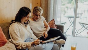 Twee personen zitten op een bank met een tablet, een glas sinaasappelsap op een tafel, en een balkon met tuinmeubilair.