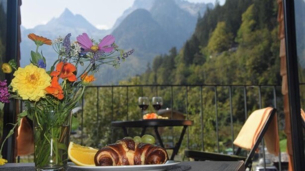 Een tafel met croissants, een glas wijn en een fruitschaal, met een berg op de achtergrond.