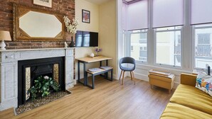 Ein Wohnzimmer mit Kamin, einem an der Wand montierten Fernseher, einem Holztisch, einem Sessel und einem Fenster mit Jalousien.