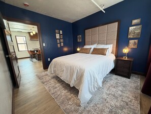 Un dormitorio con una cama grande, una mesita de noche con una lámpara y vistas a la cocina y el comedor.
