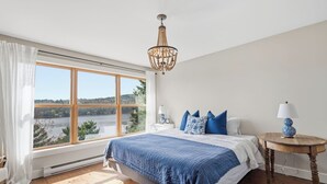 Une chambre avec une grande fenêtre, un lit avec une literie bleue, un plancher en bois et un lustre.
