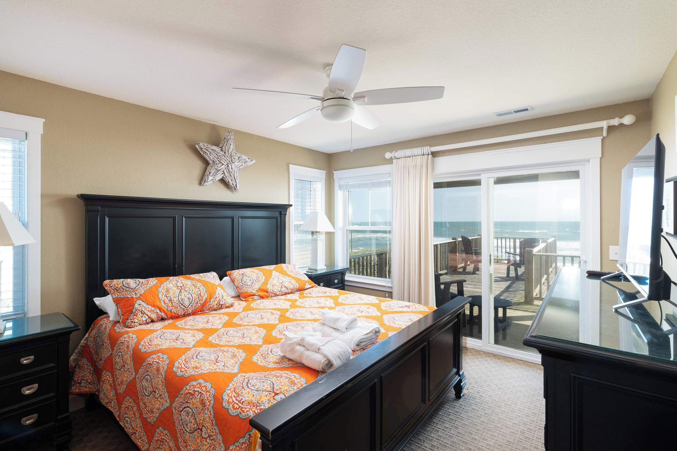 A hotel room with a bed, a ceiling fan, a star decoration, a balcony with a view, and a desk with a TV.