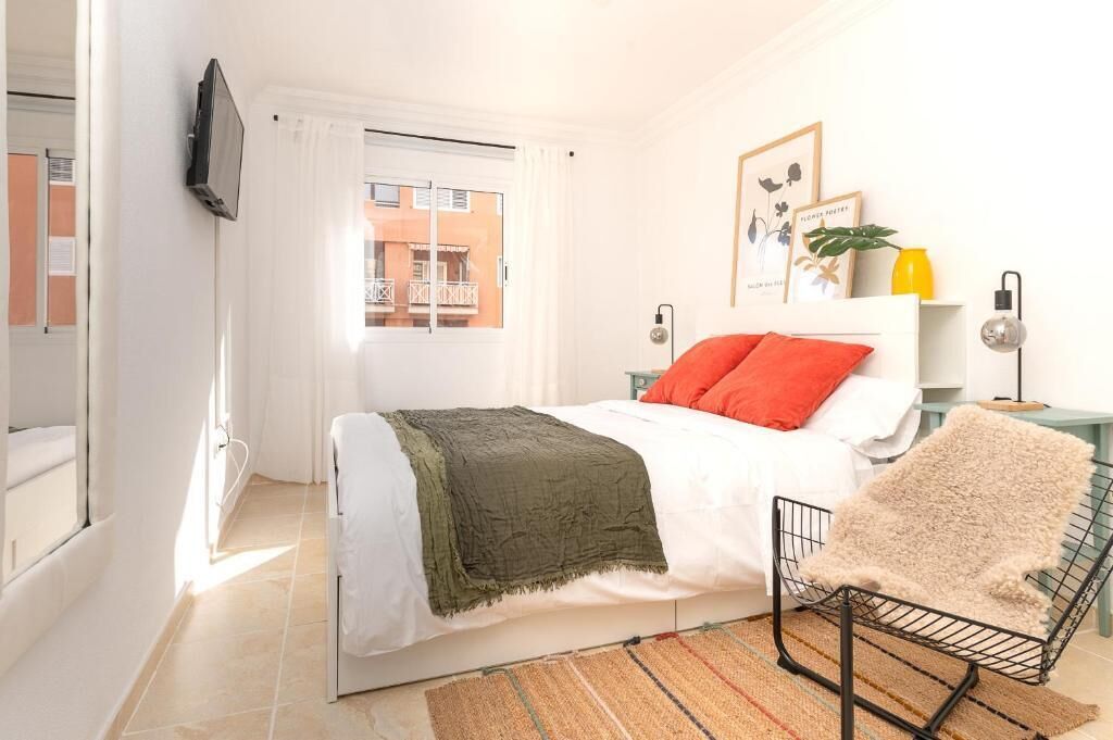 Un dormitorio con cama, una silla, televisor, ventana con cortinas, mesita de noche con lámpara y un cuadro en la pared.