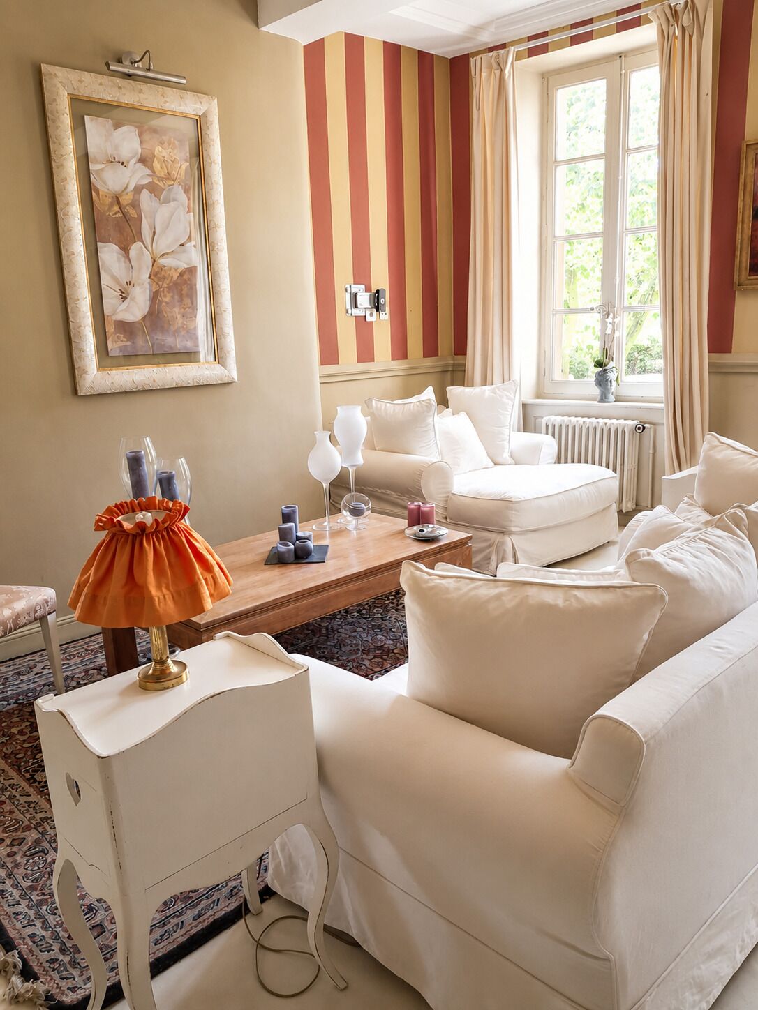 Una habitación con pared a rayas, un sofá blanco, una mesa con velas y una ventana con cortinas.
