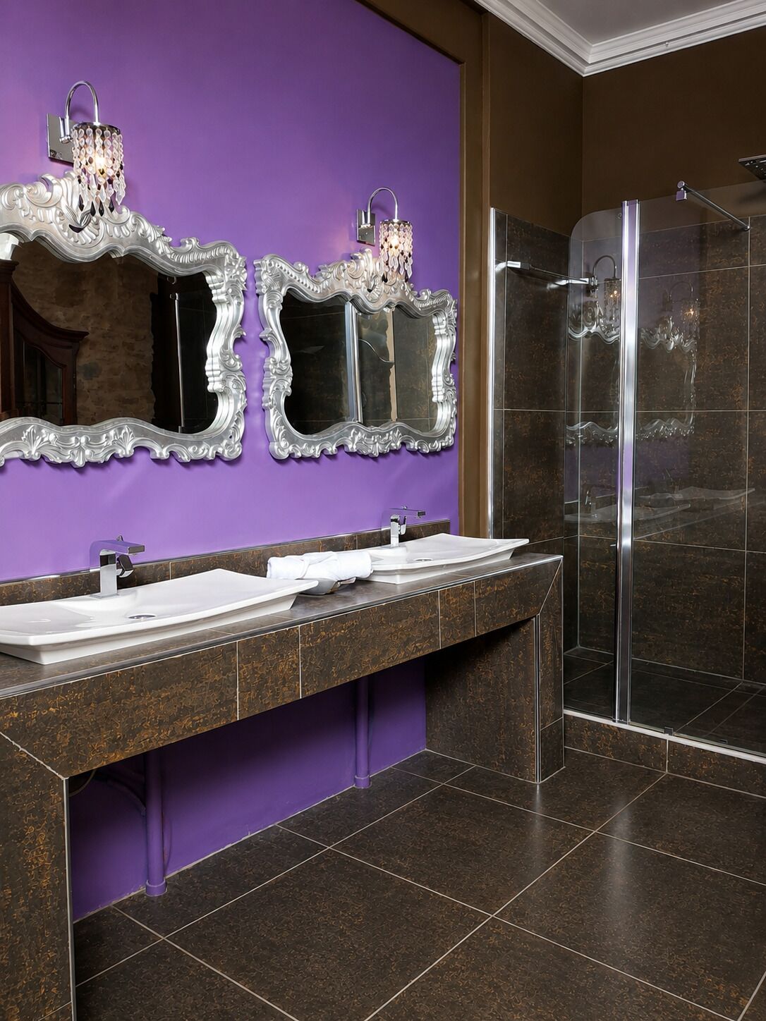 Un baño con una pared morada, dos espejos decorados, un lavabo blanco y un box de ducha de cristal.