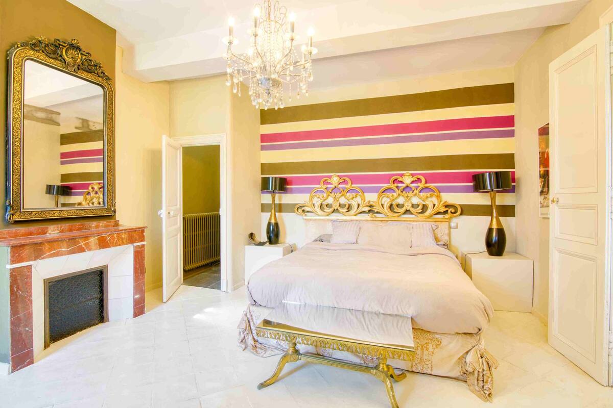 A bedroom with a large bed, a chandelier, a fireplace, and a striped wall.