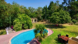 Ein Schwimmbad, umgeben von einer gepflegten Rasenfläche, einer Terrassenanlage mit Tisch und Stühlen sowie ein kleines Gebäude im Hintergrund.