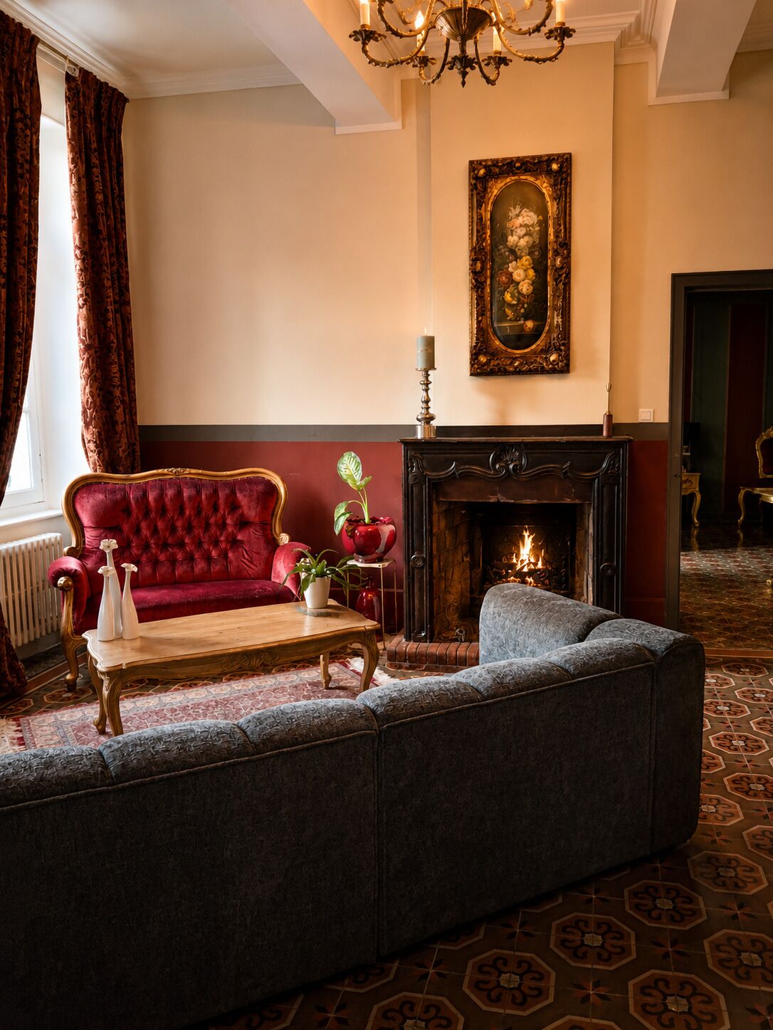 Un salon traditionnel avec une cheminée, un canapé rouge, un fauteuil gris et un lustre.