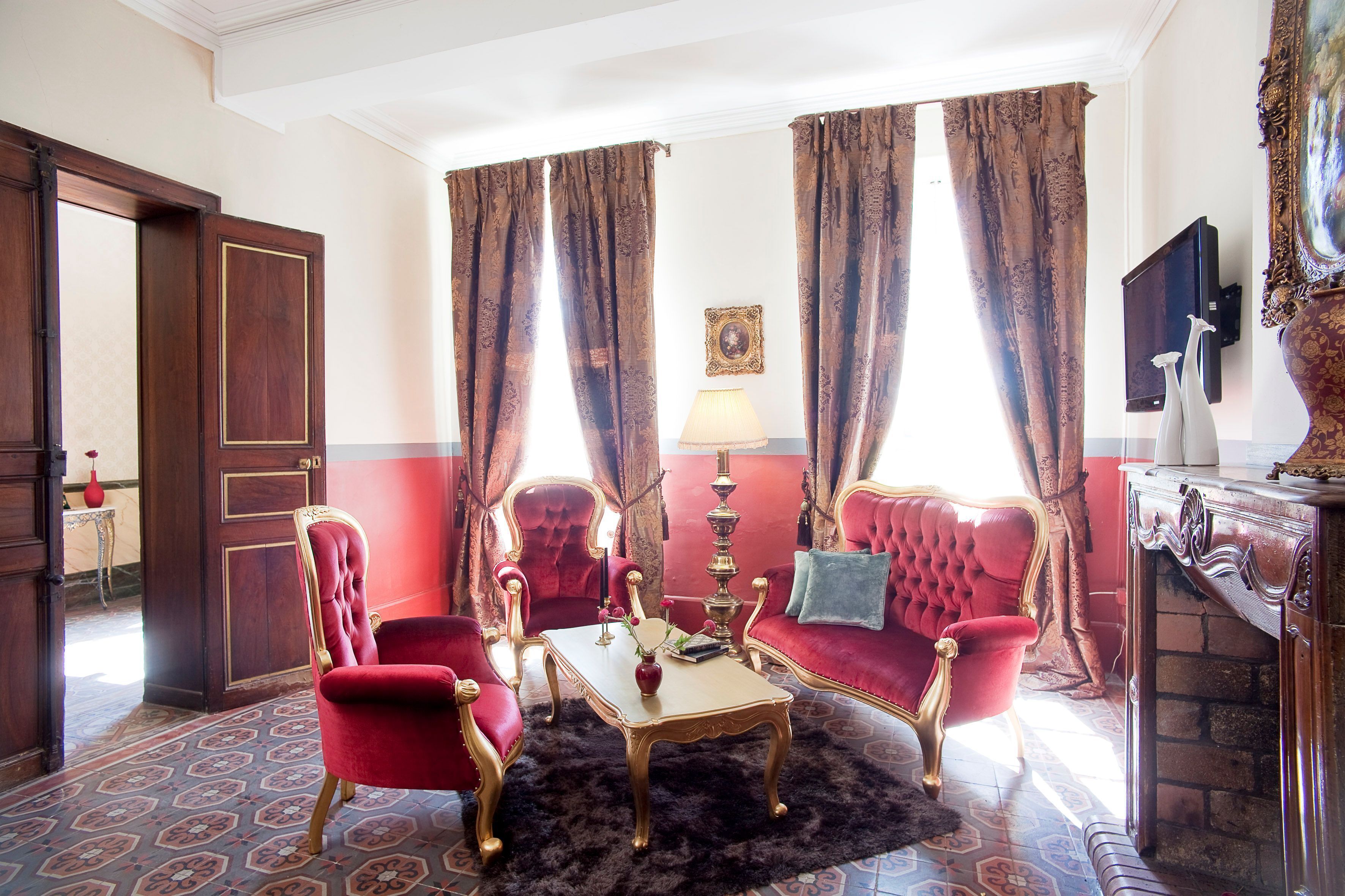 Une pièce avec un canapé rouge, deux fauteuils rouges, une petite table sur laquelle se trouve un vase, une lampe et un téléviseur fixé au mur.