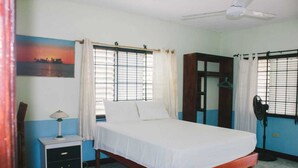 A bedroom with a bed, a nightstand, a fan, a window with curtains, and a painting on the wall. - Grassroots Negril (Negril)