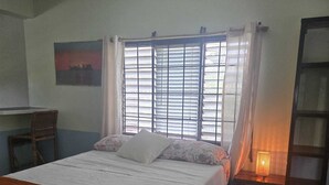 A bedroom with a bed, a desk, a chair, a window with blinds, and a nightstand with a lamp. - Grassroots Negril (Negril)