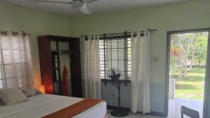 A bedroom with a bed, a ceiling fan, a window with curtains, a fan on the floor, and a view of the outdoors. - Grassroots Negril (Negril)