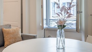 Huoneessa on pyöreä valkoinen pöytä, harmaa sohva, jossa on beigenvärinen koristetyyny, ja ikkuna, josta avautuu näkymä rakennuksiin.