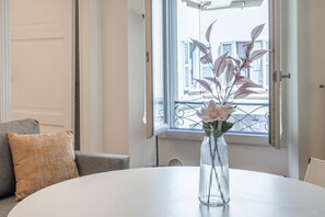 Ein Zimmer mit einem runden weißen Tisch, einem grauen Sofa mit einem beigen Kissen und einem Fenster mit Blick auf Gebäude.