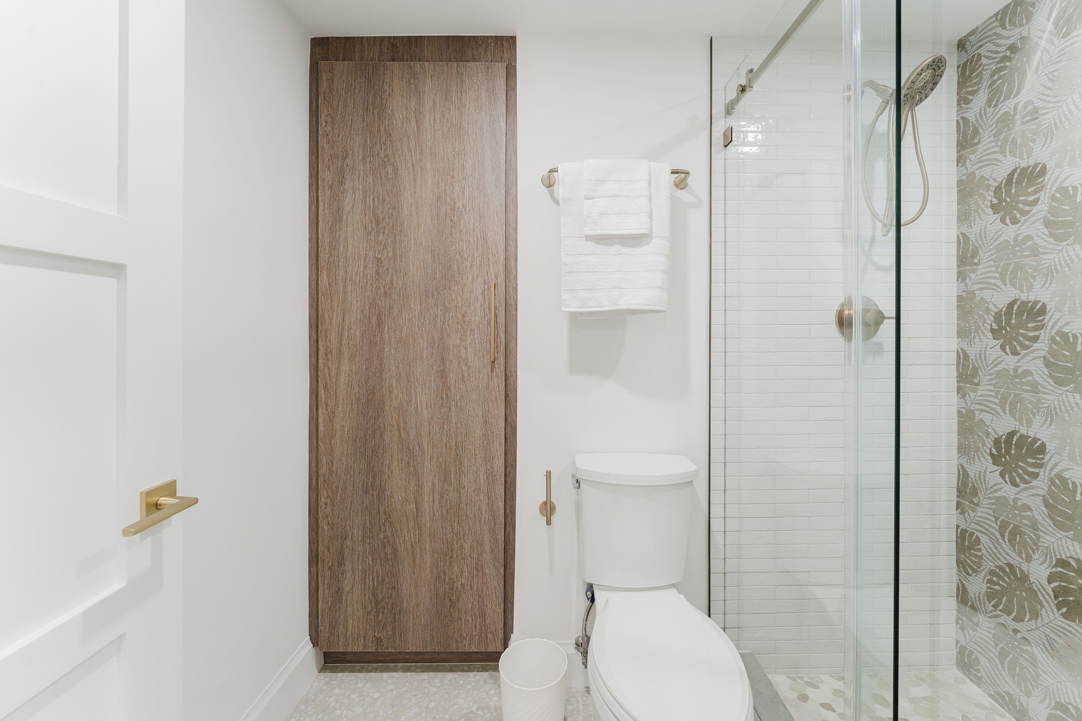 Une salle de bain avec une porte en bois, des toilettes, une douche en verre et un mur décoré d’un motif.