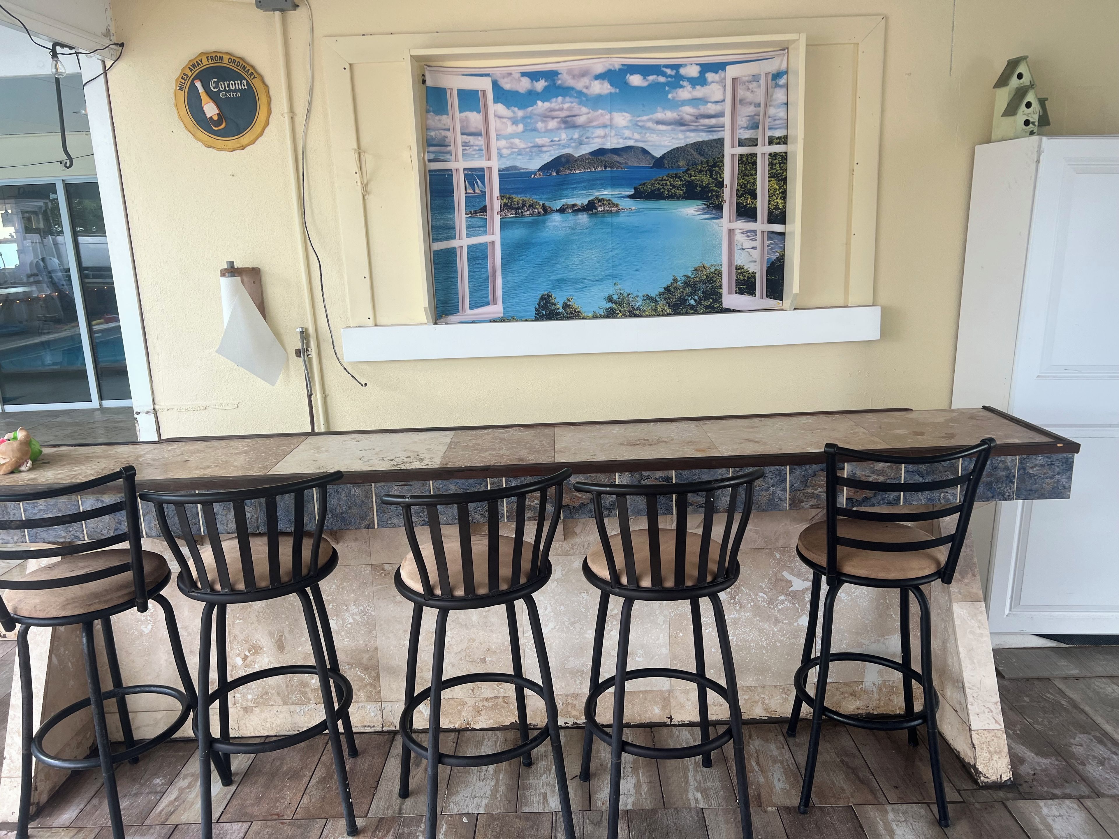 A kitchen with a long counter, four bar stools, a window with a tropical view, and a wall-mounted light fixture.