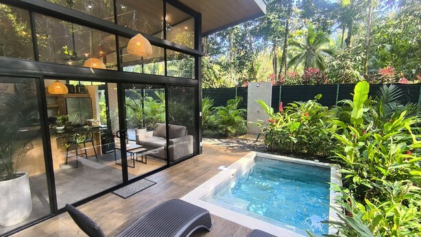 Une maison moderne à l’extérieur en verre, dotée d’une piscine et entourée d’une végétation luxuriante.