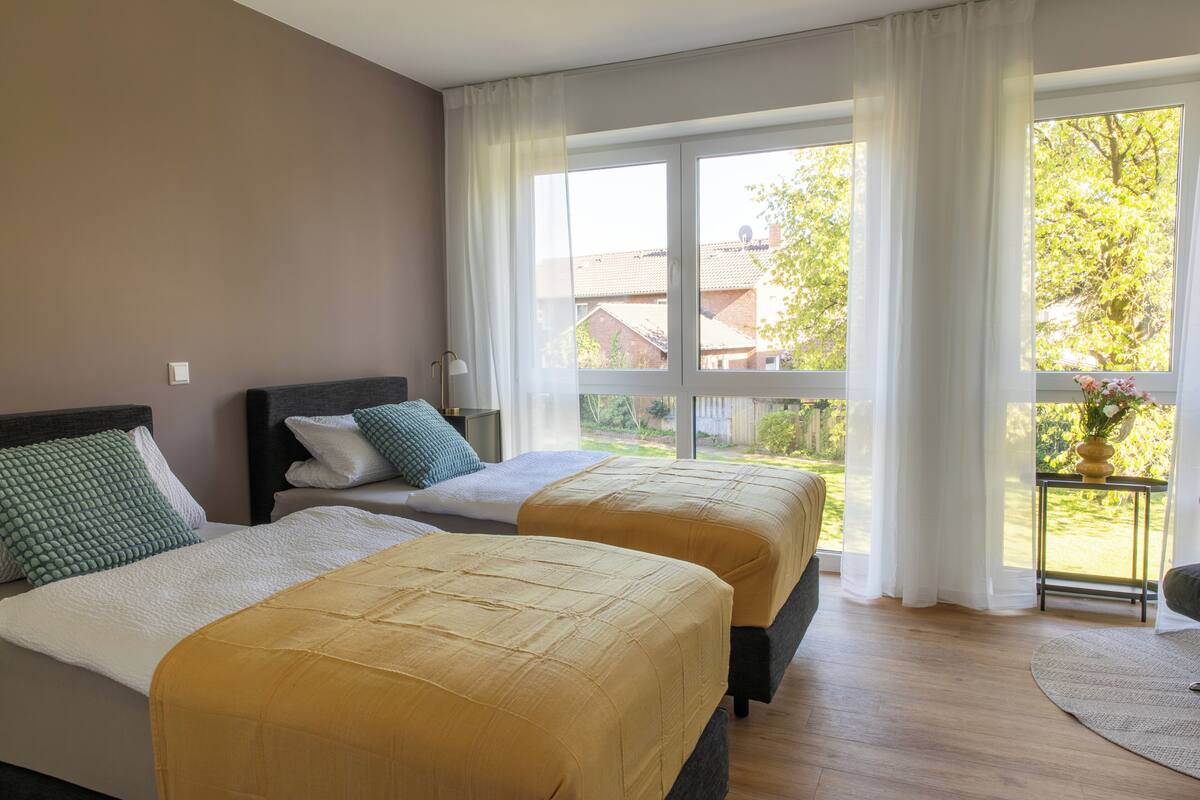 Habitación de hotel con dos camas, una ventana con cortinas, vistas al exterior y una mesita con un florero.